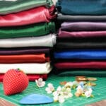 Stacks of colorful folded fabric sit on a table beside a red pincushion with pins, artificial flowers, sewing chalk, and a green cutting mat, suggesting a sewing or craft workspace.