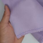 A hand holds a piece of light purple fabric with a frayed edge and visible blue stitching near the border.