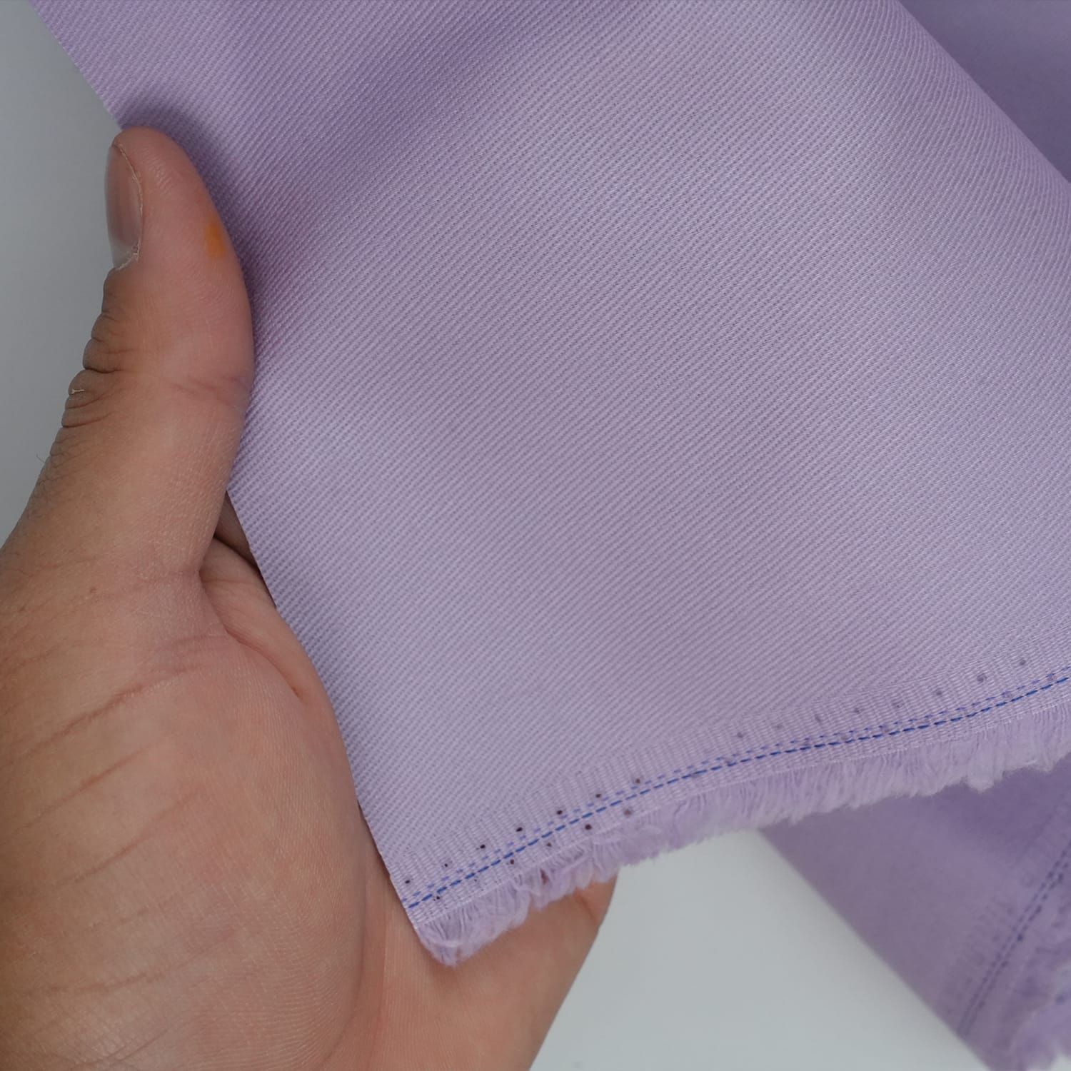 Cotton-Drill-Lilac-4 A hand holds a piece of light purple fabric with a frayed edge and visible blue stitching near the border.