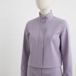 A mannequin dressed in a light purple, long-sleeved, collarless jacket with a hidden front closure and matching high-waisted pants, set against a plain white background.