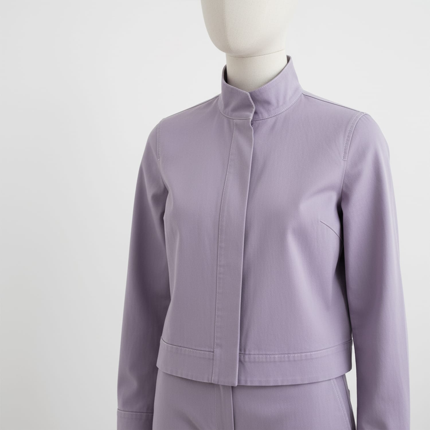 Cotton-Drill-Lilac-5 A mannequin dressed in a light purple, long-sleeved, collarless jacket with a hidden front closure and matching high-waisted pants, set against a plain white background.
