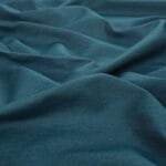 A close-up view of teal blue fabric with soft, natural folds and a slightly textured surface. The material appears smooth and lightweight, gently draped to create subtle waves and shadows.
