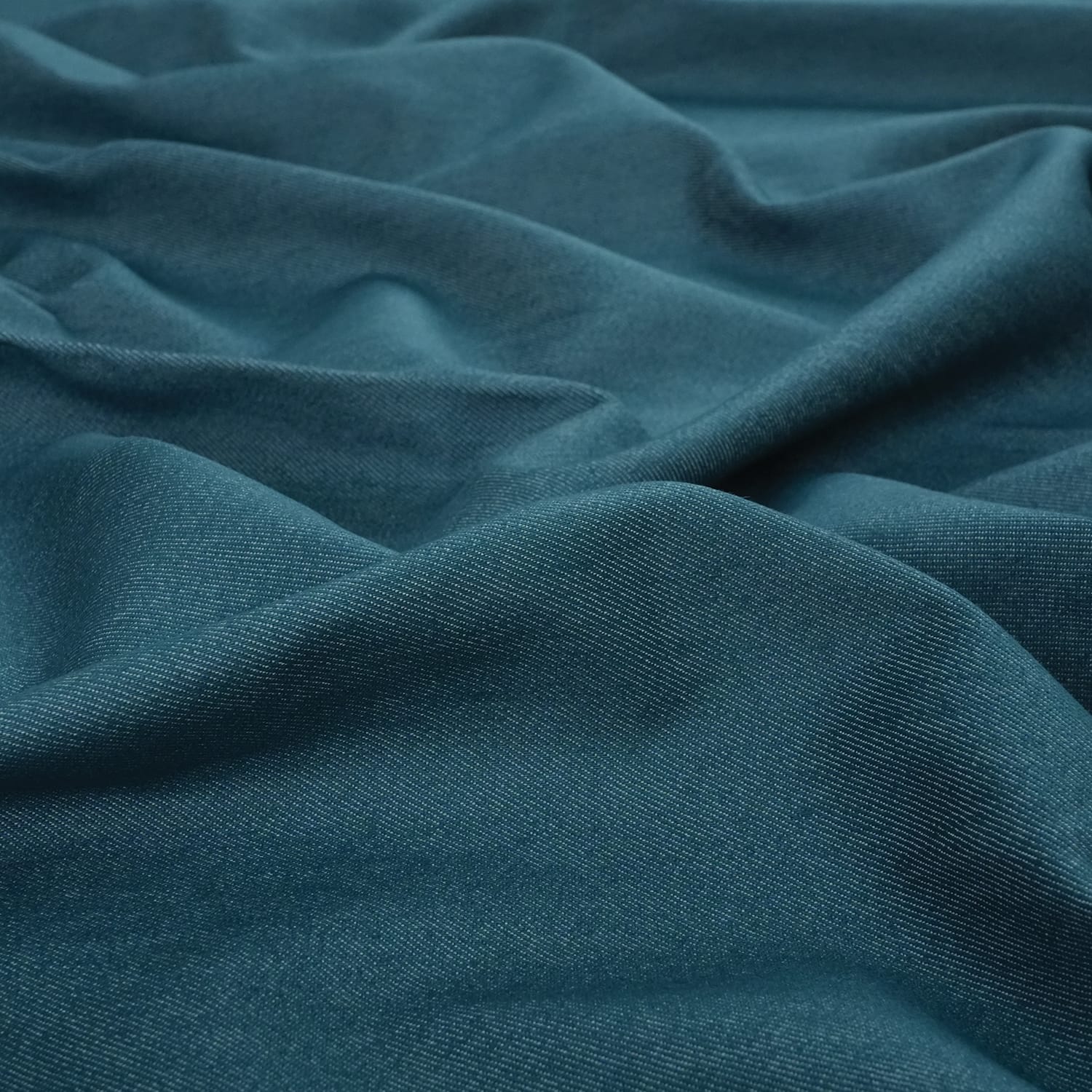 Jeans-Denim-Indigo-Petrol-4 A close-up view of teal blue fabric with soft, natural folds and a slightly textured surface. The material appears smooth and lightweight, gently draped to create subtle waves and shadows.