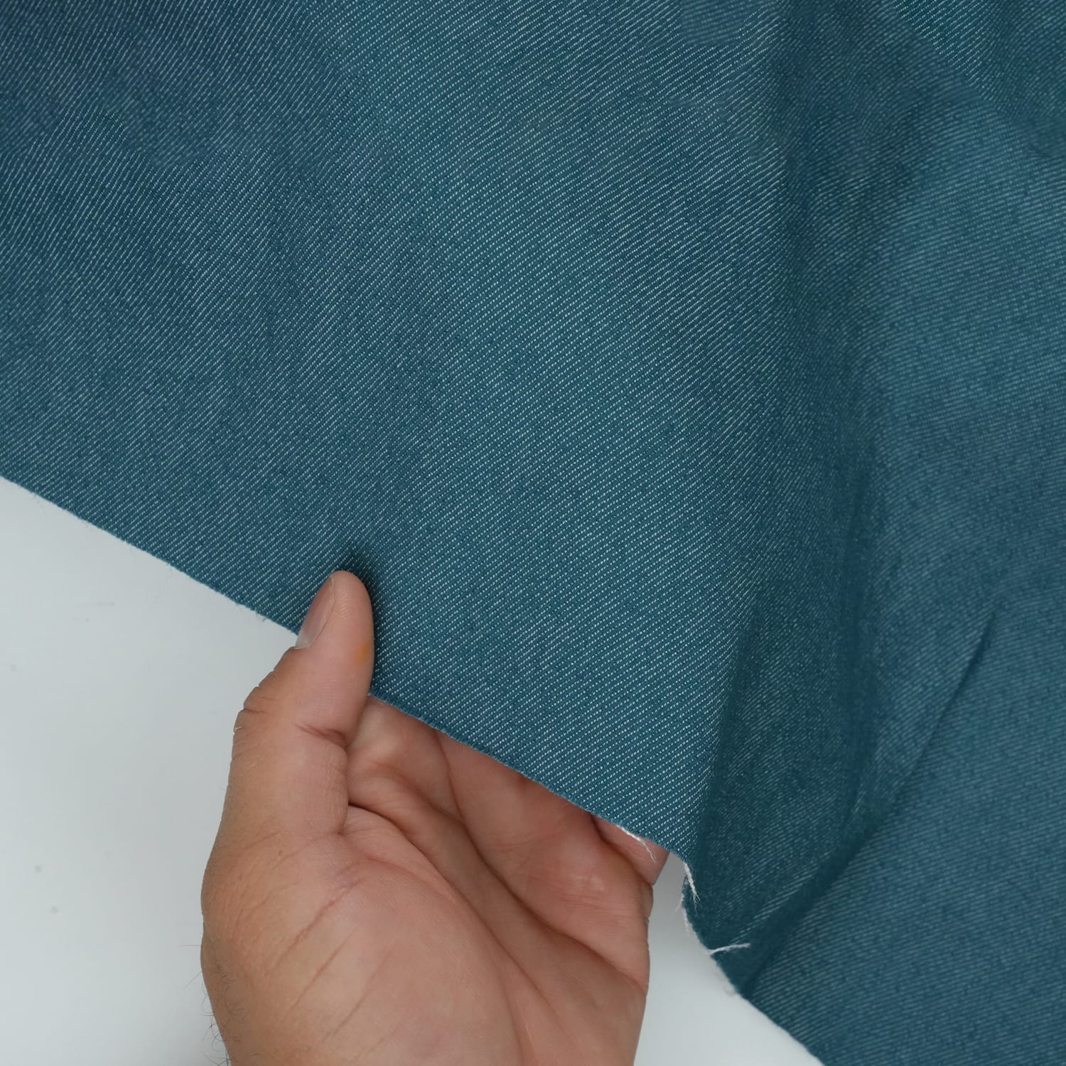 Jeans-Denim-Indigo-Petrol-5 A hand holding the edge of a piece of blue fabric, showing its texture and thickness against a white background.