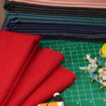 A stack of folded fabrics in various colors, red fabric laid out, spools of thread, artificial flowers, and a green cutting mat with a grid for measuring and cutting, used for sewing or crafting.