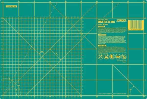 A green OLFA cutting mat marked with yellow grid lines, measurement scales in centimeters, angled guides, and warning instructions, designed for precision cutting and crafting.