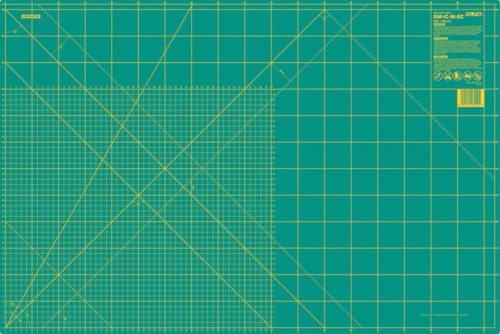 A large green self-healing cutting mat with yellow grid lines, measurement markings in inches, and angled guide lines. A label with product information is in the top right corner.