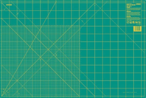 A large green self-healing cutting mat with yellow grid lines, measurement markings in inches, and angled guide lines. A label with product information is in the top right corner.