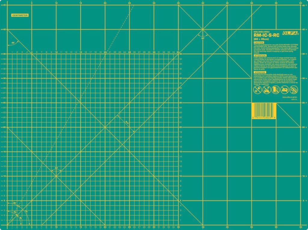 A green self-healing cutting mat with yellow grid lines, measurement marks in inches, angle guides, a barcode, safety icons, and product information in the top right corner.