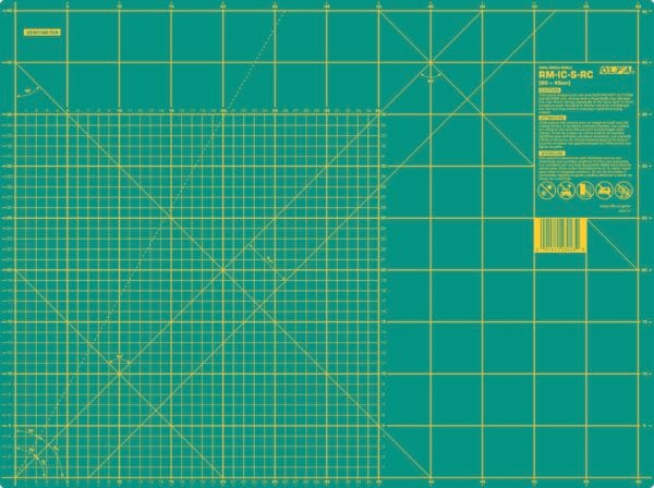 A green self-healing cutting mat with yellow grid lines, measurement marks in inches, angle guides, a barcode, safety icons, and product information in the top right corner.