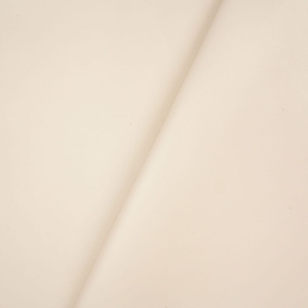 Close-up of Amigo 100% PVC Leatherette in smooth, light beige with a subtle diagonal fold from top right to bottom left. The surface is soft, uniform, and features minimal grain or markings.