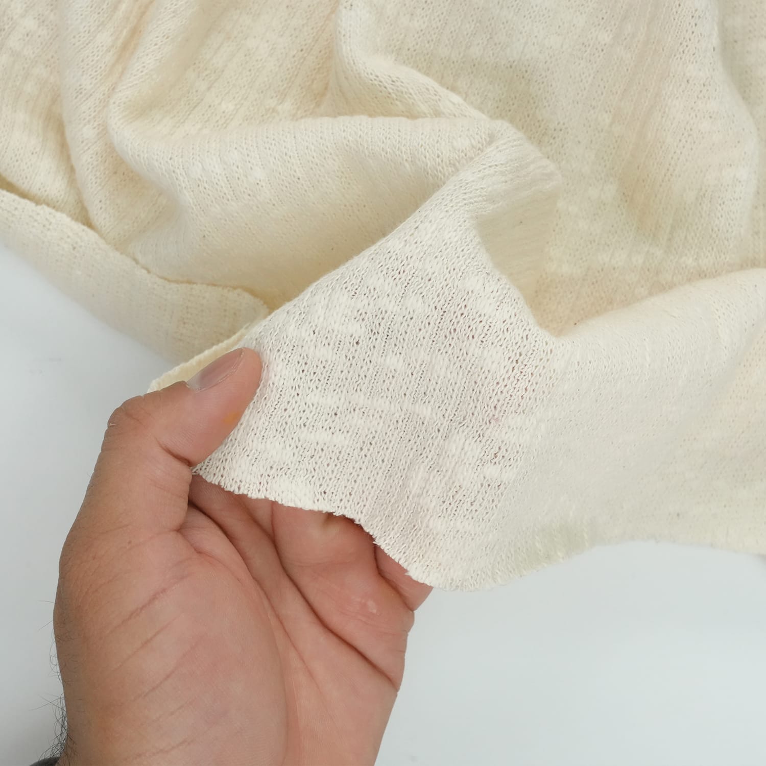 Cotton-Knitted-Cable-5 A hand holds a piece of off-white, loosely woven fabric with a textured surface, showing its softness and flexibility against a plain background.