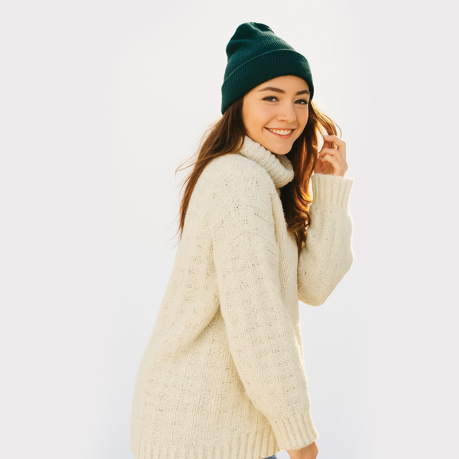Cotton-Knitted-Cable-6 A young woman wearing a cream-colored knit sweater and a dark green beanie smiles while looking over her shoulder against a light background.