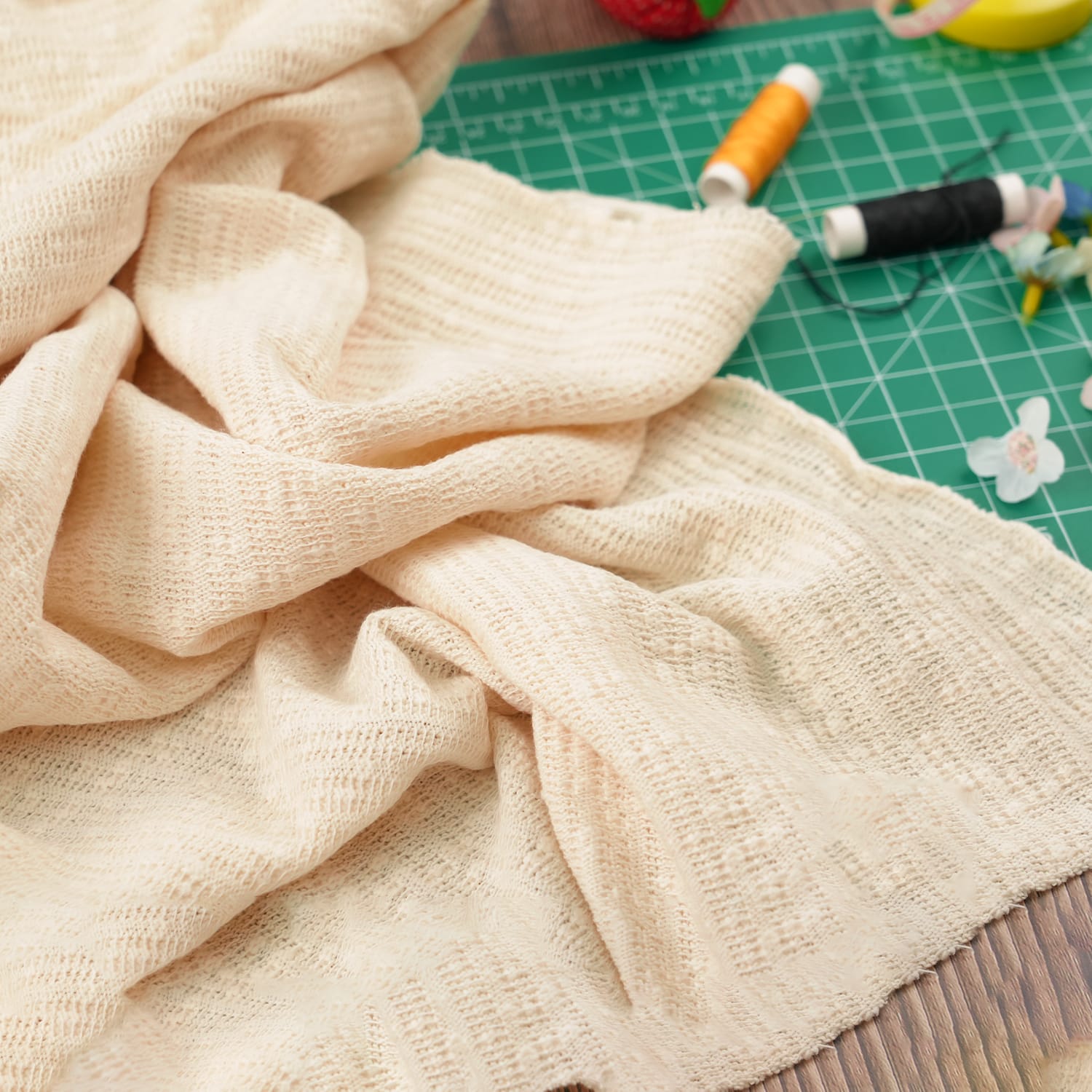 Cotton-Knitted-Cable-Variation A piece of light beige knitted fabric lies on a green cutting mat, with orange and black spools of thread, a yellow measuring tape, and small flower decorations nearby.