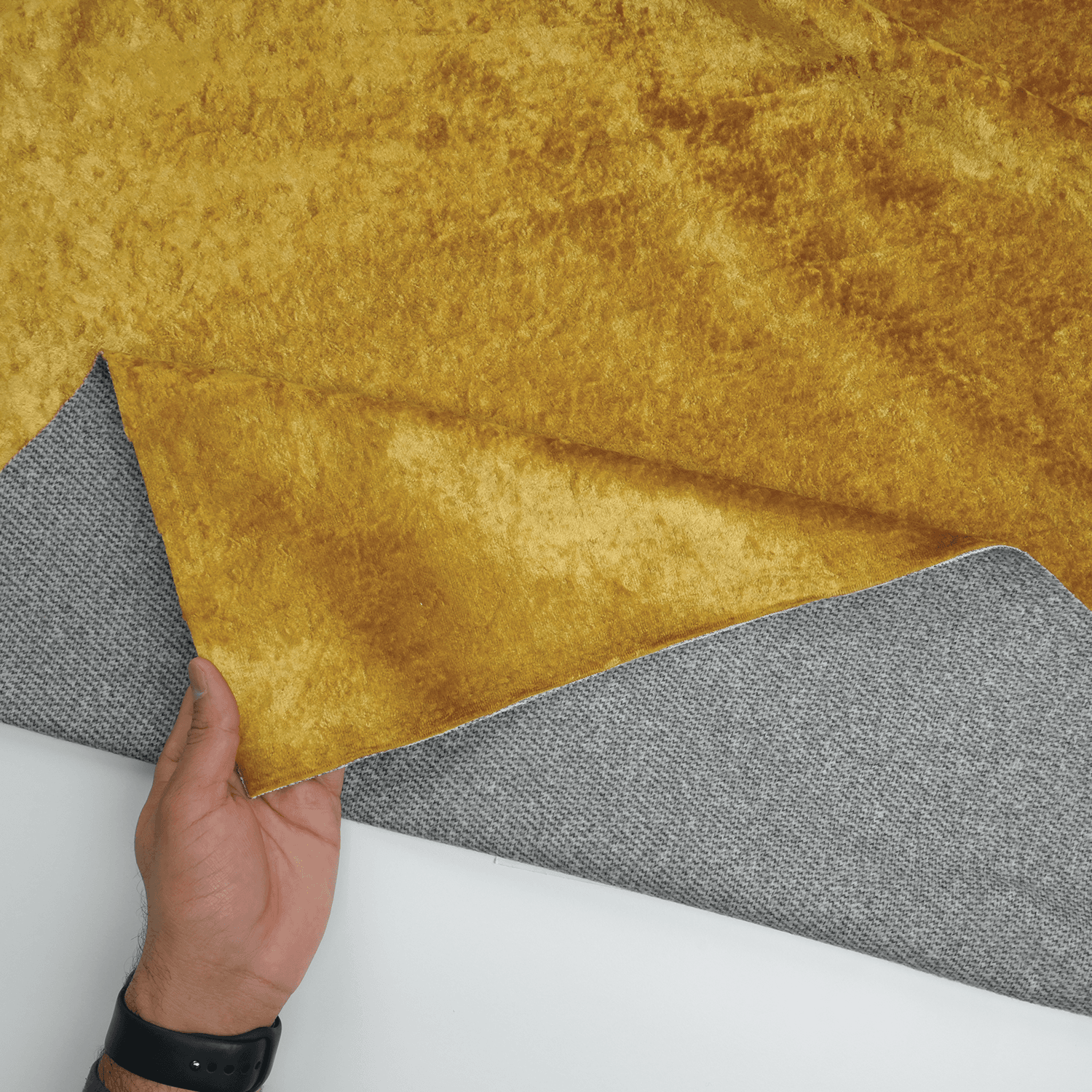 Carnival-Honey-4 A hand lifts a piece of golden-yellow velvet fabric to reveal a gray textured fabric underneath, both placed on a white surface.