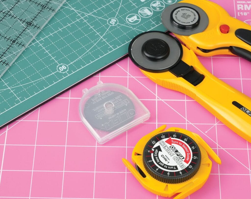 A pink cutting mat, a green gridded ruler, two yellow rotary cutters, and a circular replacement blade case are arranged on a tabletop, tools commonly used for quilting or fabric crafting.