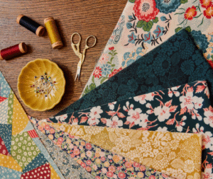 A collection of colorful patterned fabric pieces, spools of thread, a pair of gold embroidery scissors, and a yellow dish with pins arranged on a wooden surface.