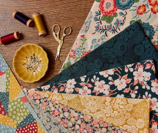 A collection of colorful patterned fabric pieces, spools of thread, a pair of gold embroidery scissors, and a yellow dish with pins arranged on a wooden surface.